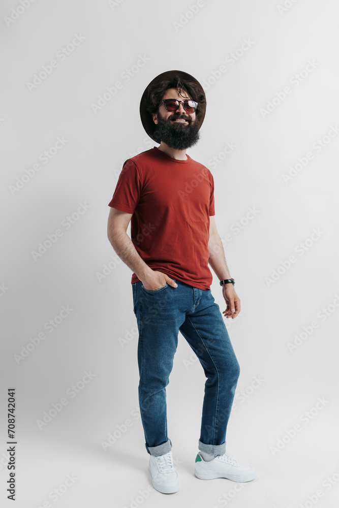 Fashionable man with a beard wearing a red tee, denim jeans, and a wide-brimmed hat, striking a pose with sunglasses