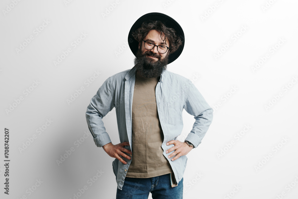 Portrait of a confident, bearded man in a hat, hands on hips, wearing casual clothes against a white backdrop Portrait of a confident, bearded man in a hat, hands on hips, wearing casual clothes against a white backdrop