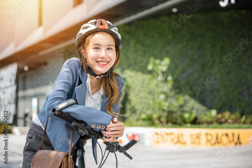 Fototapeta Naklejka Na Ścianę i Meble -  In the city an Asian businesswoman helmeted and in a suit stands with her bicycle ready for a cheerful morning commute to the office. This image combines work and outdoor fun.