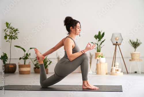 Side view of young caucasian girl in grey sportswear doing yoga. One-legged posture King Pigeon pose or Eka Pada Rajakapotasana. Calm woman controlling breath and meditating with eyes closed at home. photo