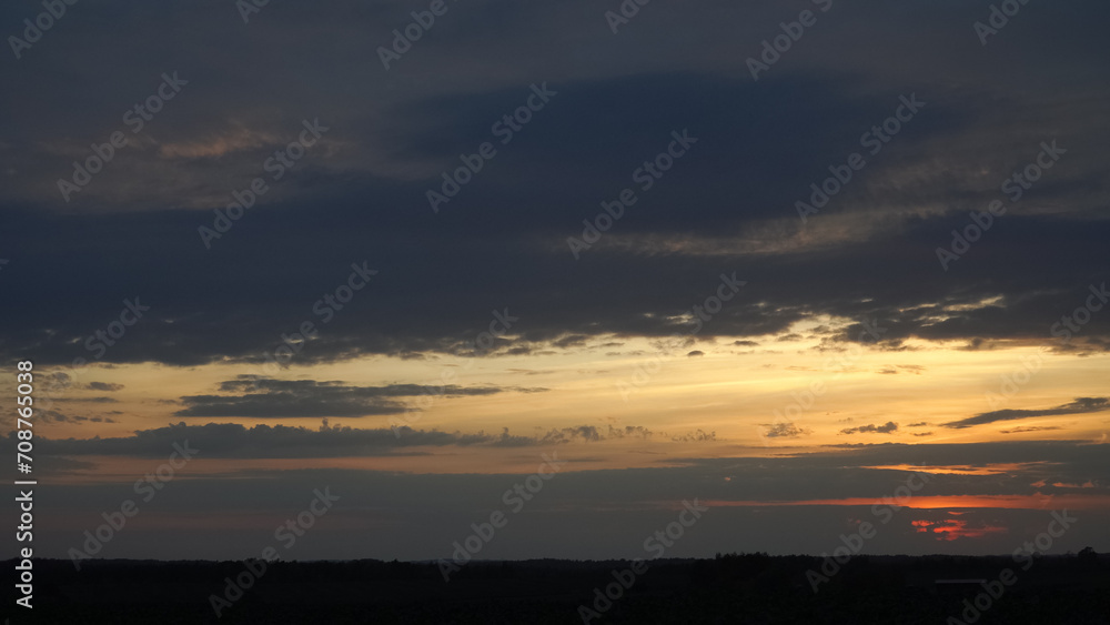 Panoramic view of a golden sunset sky with dramatic, dark grey clouds. The beautiful evening landscape showcases a visible horizon line on a plain at nightfall.