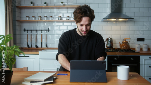 Happy curly hair man sitting in kitchen, using app with tablet computer, surfing on internet 