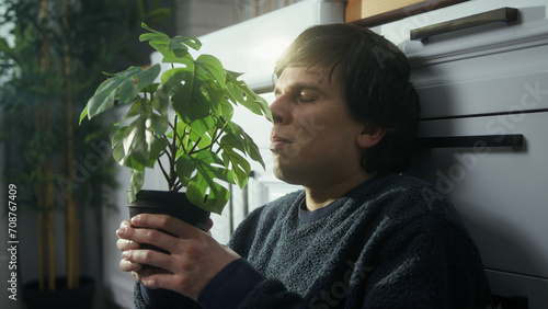 Eyes closed man sitting on the floor in the kitchen showing love and smells the flower in the pot he holds in his hand	
