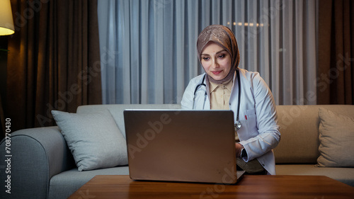 Female doctor in headscarf wears white coat talking to patient using virtual chat computer app while sitting on sofa 