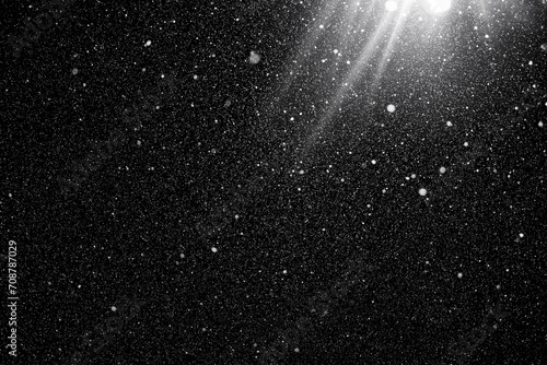 Floating dust particles. White dust texture on a black background with a bright ray of light coming through. Snowflakes falling at night from the sky. 