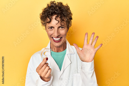 Doctor with invisible dental aligner on yellow smiling cheerful showing number five with fingers.