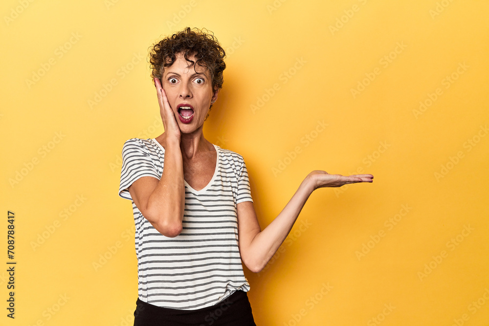 Mid-aged caucasian woman on vibrant yellow holds copy space on a palm, keep hand over cheek. Amazed and delighted. - obrazy, fototapety, plakaty 
