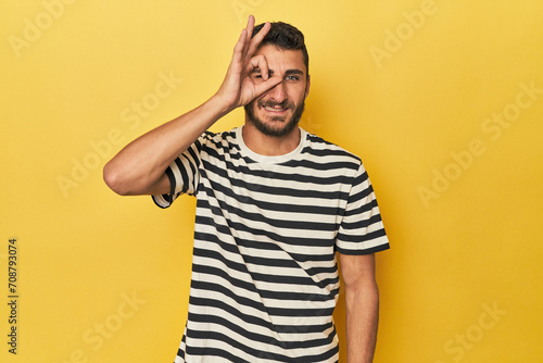 Young Hispanic man on yellow background excited keeping ok gesture on eye.