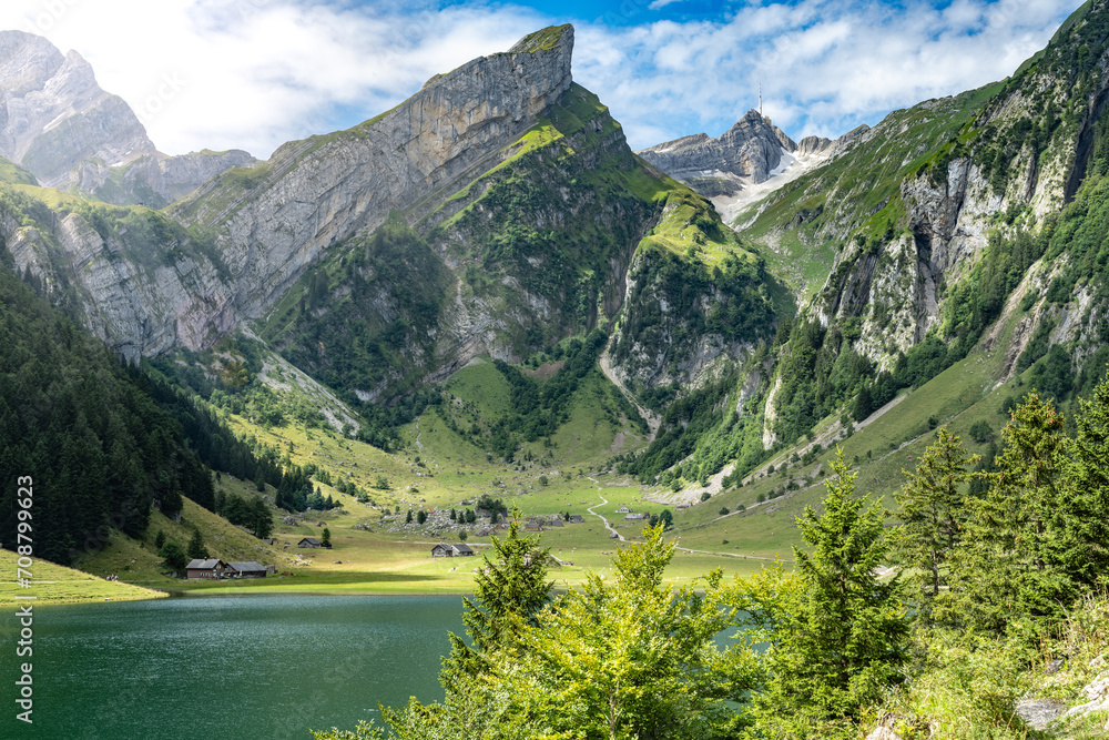 Panoramic view of an alpine lake in a green valley with mountain huts and mountain peak in the background on a sunny day. Seealpsee, Säntis, Wasserauen, Appenzell, Switzerland.