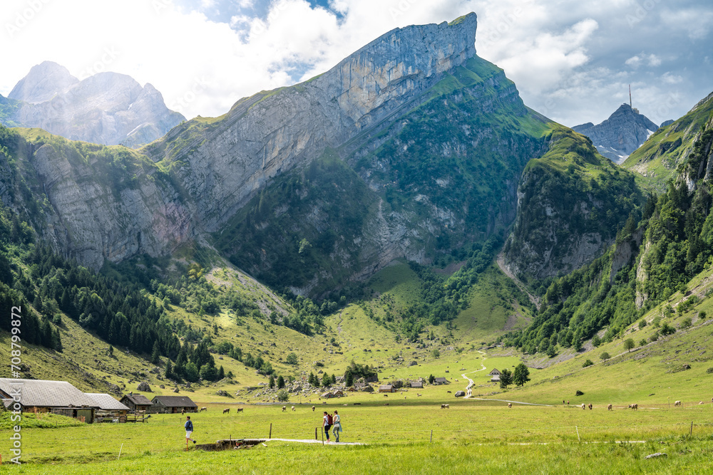 Panoramic view of tourists walking along trail in a green valley with a mountain peak in the background on a sunny day. Seealpsee, Säntis, Wasserauen, Appenzell, Switzerland.