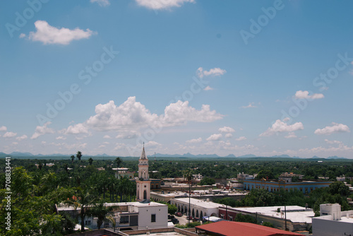 A picturesque town with a prominent church spire, surrounded by lush greenery and a mountain range under a cloud-speckled sky.