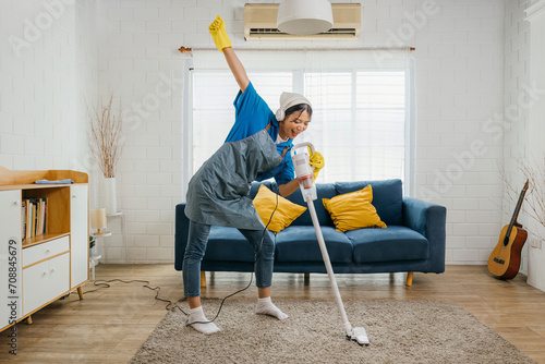 Happy young Asian housewife sings her favorite song with a vacuum cleaner as a microphone while cleaning. Enjoying domestic work dancing in living room a cheerful maid having fun.