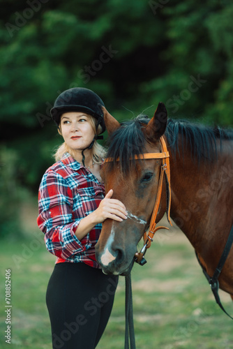 Happy blonde with horse in forest. Woman and a horse walking through the field during the day. Dressed in a plaid shirt and black leggings.