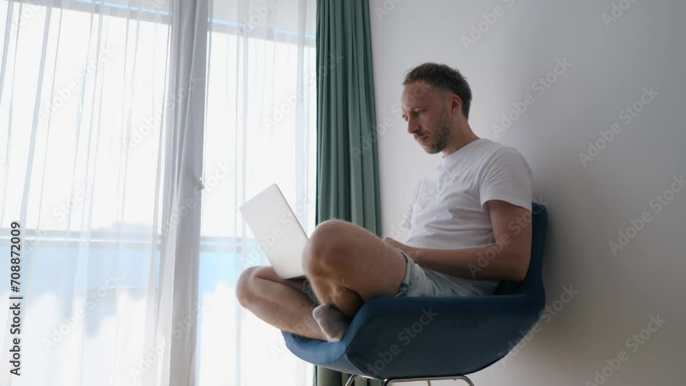Man works at laptop sitting on chair and cross-legged. Freelancer or remote worker using computer at home Man works at laptop sitting on chair and cross-legged. Freelancer or remote worker using computer at home