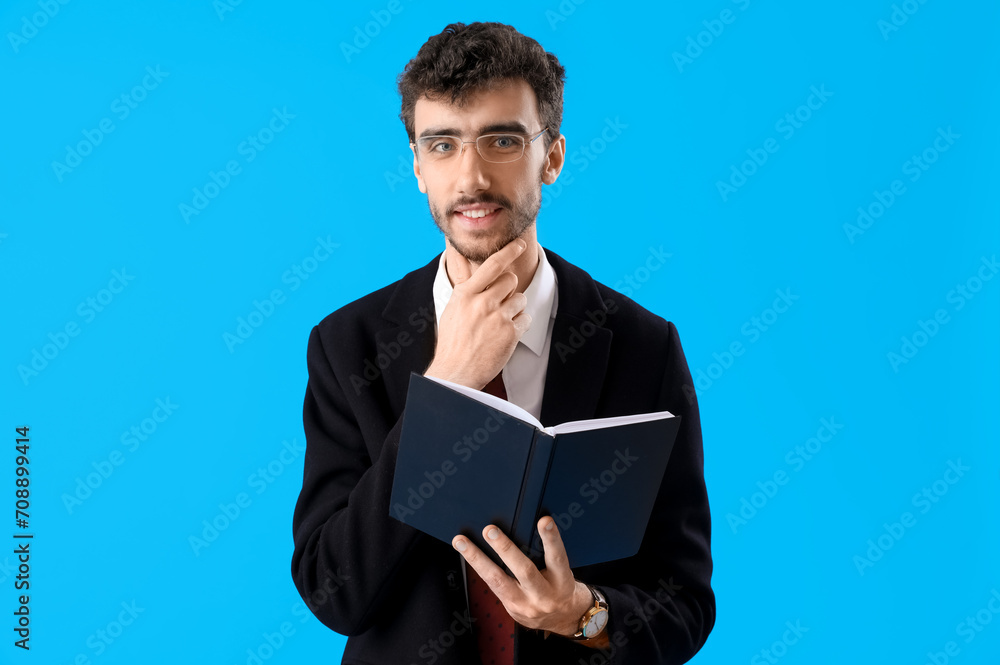 Thoughtful young man in suit with notebook on blue background Thoughtful young man in suit with notebook on blue background