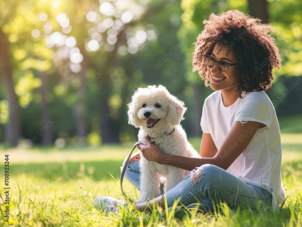 African american woman with curly hair and eyeglasses sitting on the grass in the park with her dog.