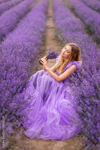 young woman in a lilac dress is sitting in a field with lavender. She has a bouquet of fragrant flowers in her hands