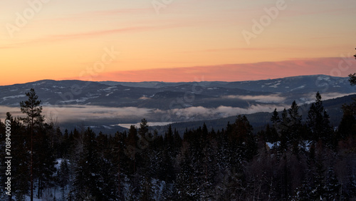 Misty landscape with foreground trees, a foggy valley, and Lake Krøderen in Norway. The serene scene is bathed in a captivating red sunrise sky. © Barnabas