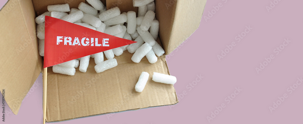 An overturned open cardboard packaging box with fallen white polystyrene packaging chips. Tiny paper flag with the warning 'Fragile' as a label. The concept of shipping fragile products - obrazy, fototapety, plakaty 