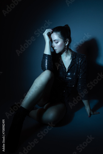 Studio fashion portrait of a beautiful Ukrainian model. She is wearing a black shiny jumper and is posing while sitting on the floor. A blue gel was used to create a blue tone. 