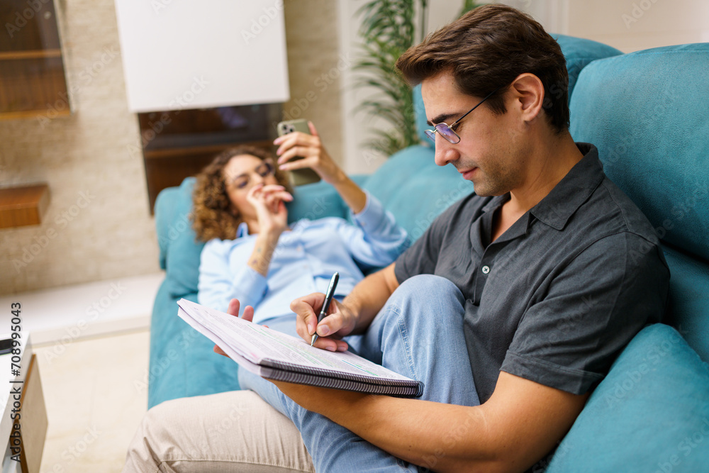 Focused young husband taking notes in notebook while sitting with wife on sofa Focused young husband taking notes in notebook while sitting with wife on sofa