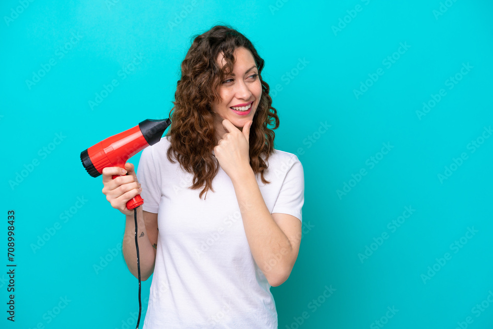 Young caucasian woman holding a hairdryer isolated on blue background thinking an idea and looking side Young caucasian woman holding a hairdryer isolated on blue background thinking an idea and looking side