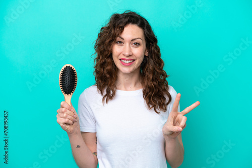 Young caucasian woman holding hairbrush isolated on blue background smiling and showing victory sign photo