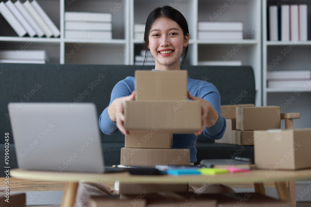 Young Asian woman working with parcel box at home preparing to deliver goods and writing online order note from internet on laptop. Young Asian woman working with parcel box at home preparing to deliver goods and writing online order note from internet on laptop.