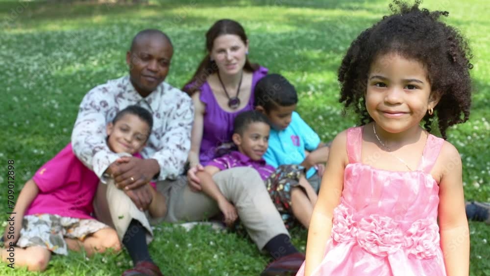 Little girl standing in park, family sitting on the grass behind Little girl standing in park, family sitting on the grass behind