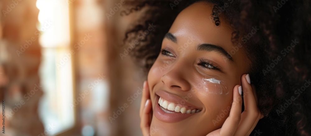 A smiling millennial woman, of African American descent, applies cream on her face at home in the bedroom mirror in the morning, using cosmetics for skin care and enjoying a spa-like experience. A smiling millennial woman, of African American descent, applies cream on her face at home in the bedroom mirror in the morning, using cosmetics for skin care and enjoying a spa-like experience.