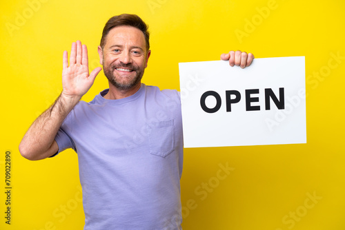 Middle age caucasian man isolated on yellow background holding a placard with text OPEN and saluting © luismolinero