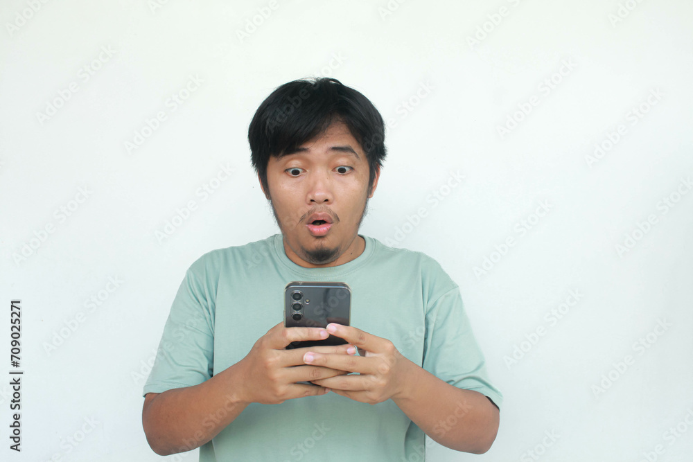 Portrait of a young Asian man in a green t-shirt casually holding his cellphone with a happy and wow expression. Isolated on gray background. Portrait of a young Asian man in a green t-shirt casually holding his cellphone with a happy and wow expression. Isolated on gray background.