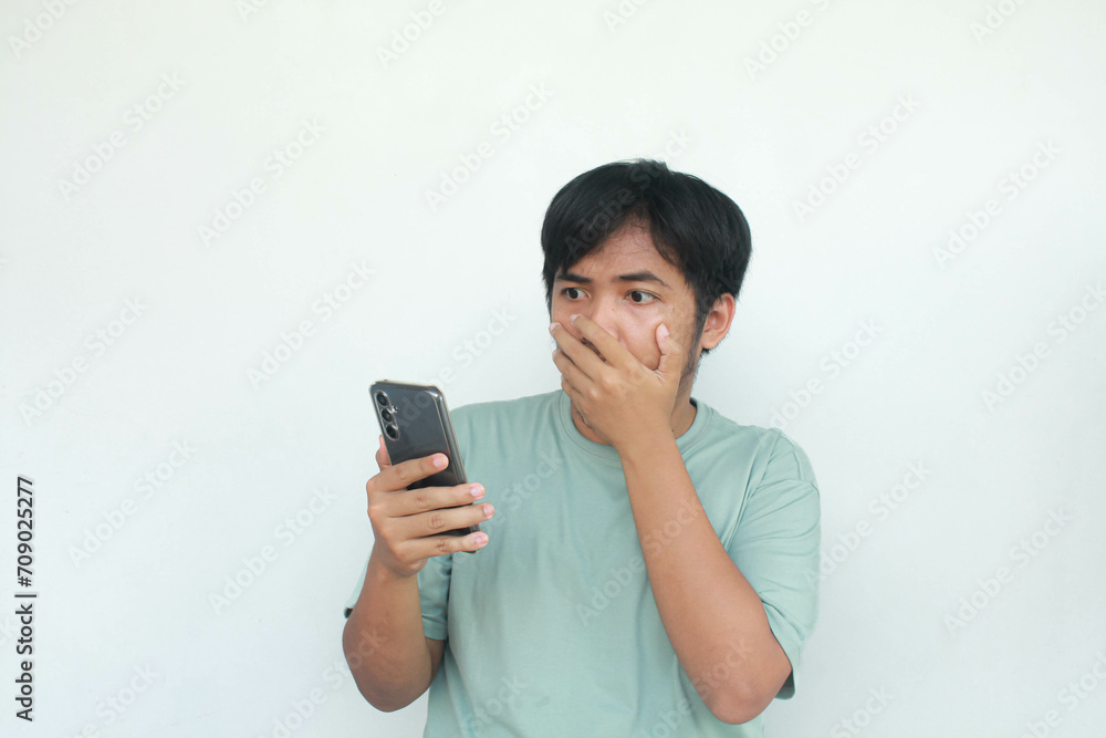 Young Asian man showing surprised expression while typing on his mobile phone. Isolated in gray background. Young Asian man showing surprised expression while typing on his mobile phone. Isolated in gray background.