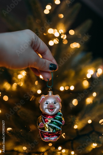 A New Year's glass toy in the shape of a pig in the girl's hand on the background of an artificial Christmas tree with a garland with warm blurred lights. The symbol of the year is a pig