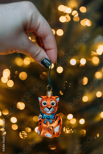 A New Year's glass toy in the shape of a tiger in the girl's hand on the background of an artificial Christmas tree with a garland with warm blurred lights. The symbol of the year is a tiger