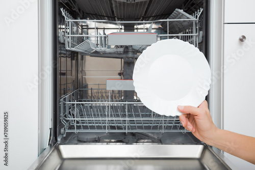 Young adult woman hand holding and showing white clean plate on dishwasher background at home kitchen. Closeup. Front view.