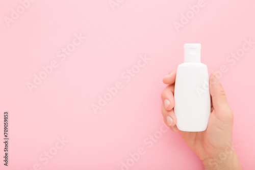 Young adult woman hand holding white plastic cream bottle on pink table background. Pastel color. Care about clean and soft body skin. Female beauty product. Closeup. Empty place for text. Top view.
