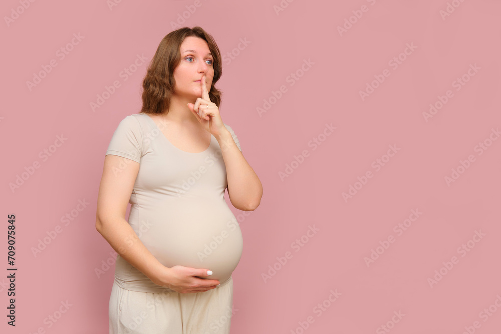 A pregnant woman with her finger on her lips on a pink studio background. Pregnancy in a woman with a belly showing a sign of silence, copy space A pregnant woman with her finger on her lips on a pink studio background. Pregnancy in a woman with a belly showing a sign of silence, copy space
