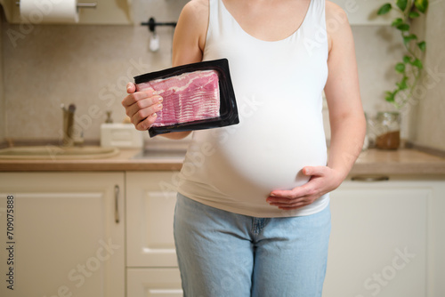 A pregnant woman holds bacon meat in her hands while standing in a home kitchen. Pregnancy and proper nutrition while waiting for a baby
