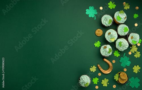 St. Patrick's Day vanilla and chocolate cupcakes with green frosting and  shiny clover decorations on green paper background. Irish holiday dessert concept. Top view, copy space.
