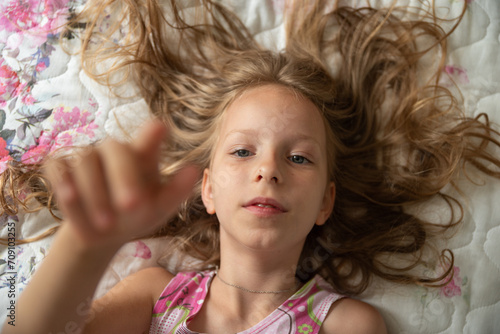 Photo of Cute girl enjoying the morning in bed, indoors at home make selfie by camera lies in bed waving.