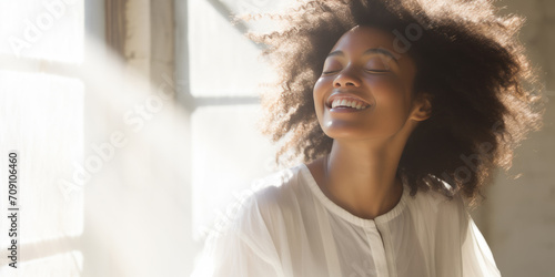 Woman smiles blissfully with closed eyes in front of a window, basking in radiant morning light.