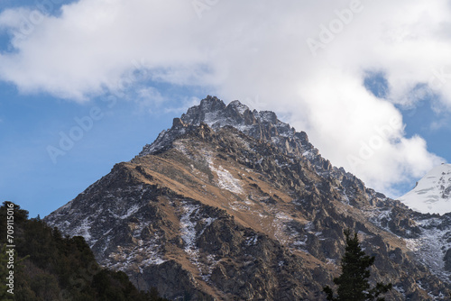 High mountains dusted with the first snow of autumn. A minimalist mountain landscape with clouds and blue sky overhead. In the distance, a glacier is visible.