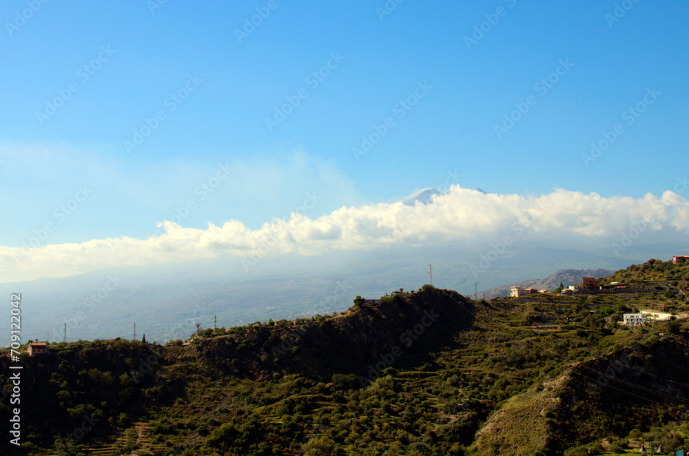 Fototapeta premium Panoramic nature landscape. Mountains against blue sky. Famous Sicilian Etna volcano in white clouds in the background. View from Taormina. Travel and tourism concept