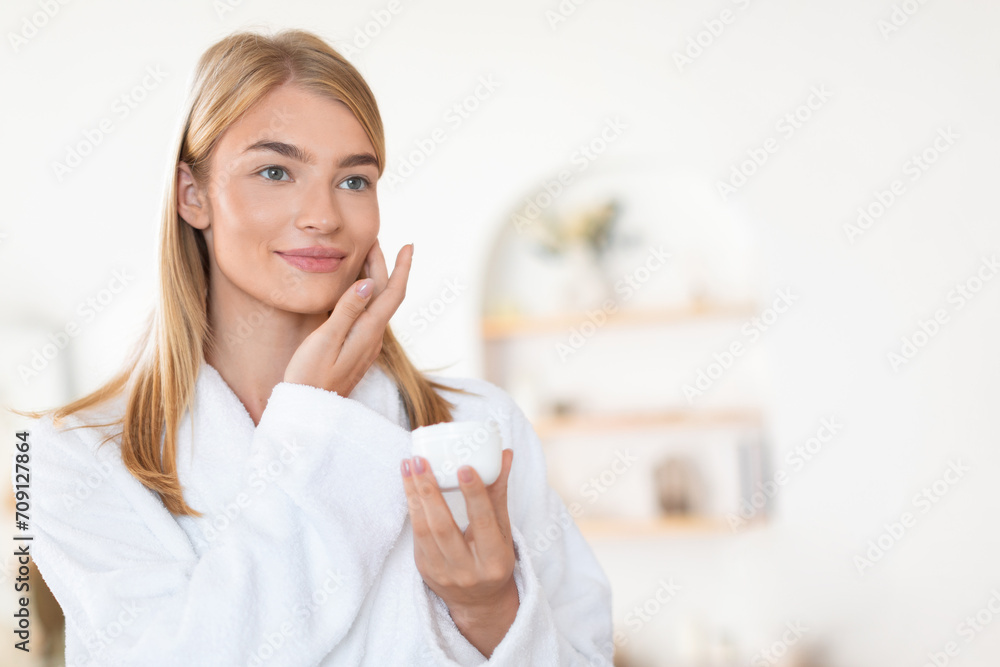 Young blonde woman in a bathrobe enjoys skin moisturizing indoor Young blonde woman in a bathrobe enjoys skin moisturizing indoor