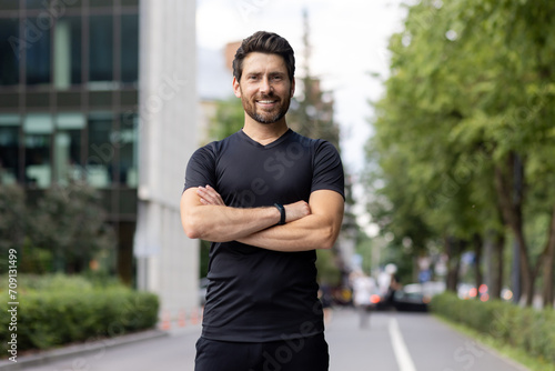 Portrait of a young sportsman standing on a city street, arms crossed on his chest, confidently and smilingly looking at the camera
