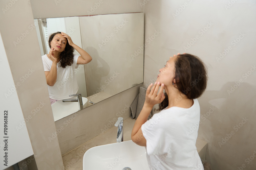 Happy young woman touching her face, examining flaws, skin problems and wrinkles, standing in front of a bathroom mirror Happy young woman touching her face, examining flaws, skin problems and wrinkles, standing in front of a bathroom mirror