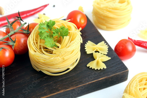 A black wooden cutting board with vegetables and pasta lies on a white background.