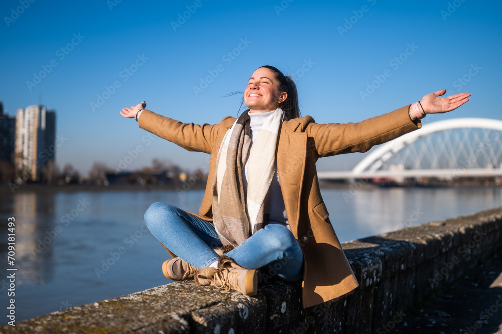 Beautiful woman in warm clothing with her arms outstretched enjoys resting by the river on a sunny winter day. Beautiful woman in warm clothing with her arms outstretched enjoys resting by the river on a sunny winter day.