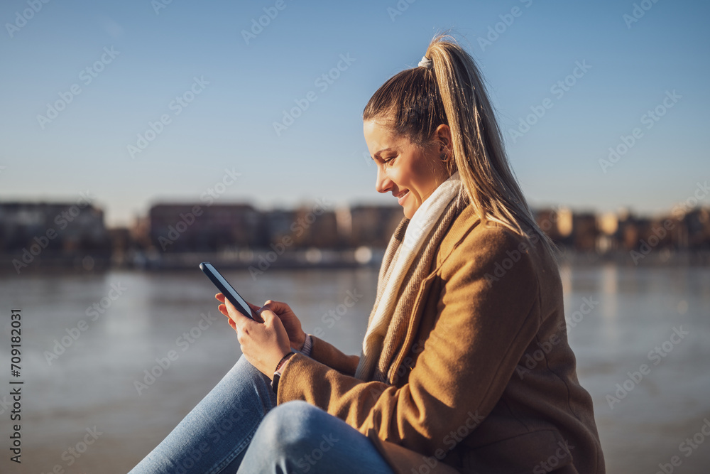 Beautiful woman in warm clothing using mobile phone and enjoys resting by the river on a sunny winter day. Toned image.